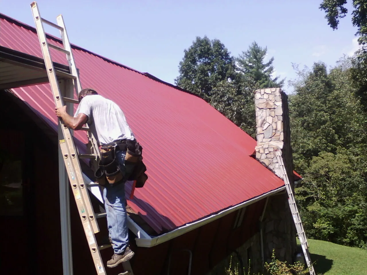 Roofer on a ladder beside a red metal roof and stone chimney — Soffit & Fascia Repair by Roofing Brothers in Hornsby